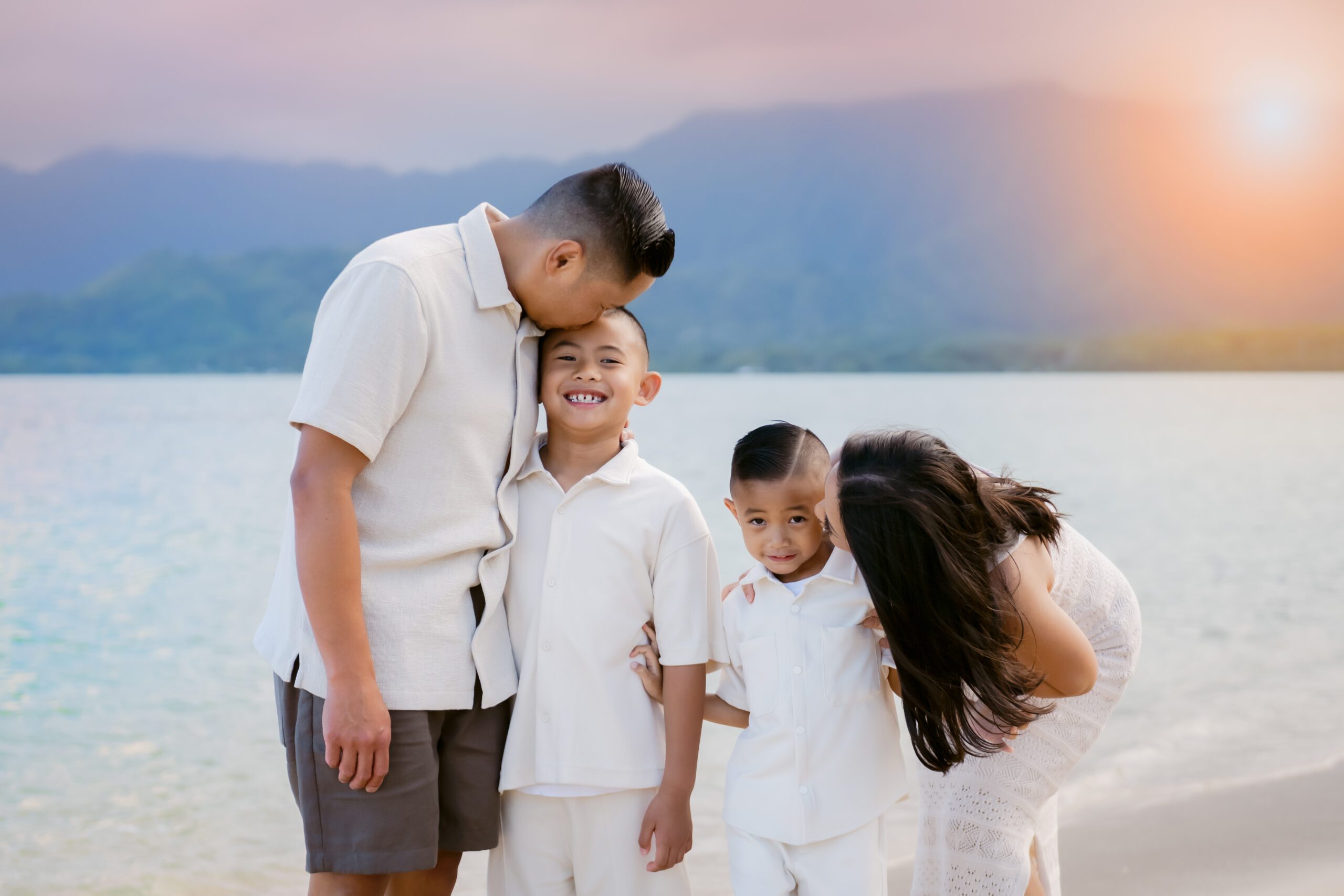 Family of four sharing a tender moment on the beach at sunset with mountains in the background.
