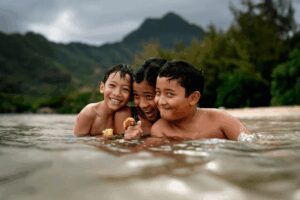 Three smiling kids play in the water, surrounded by mountains and greenery.